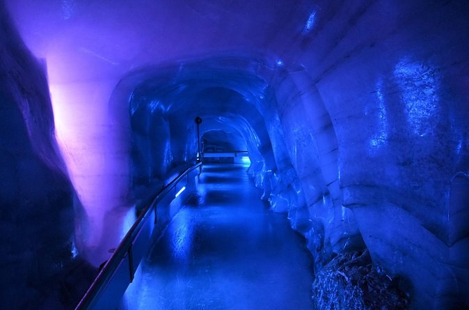 A tunnel through glacier ice lit deep blue, with a handrail along its wall